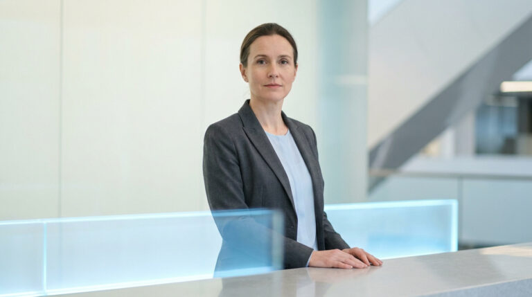 Une femme d'affaires sérieuse en costume gris derrière un comptoir d'accueil dans un bureau moderne et minimaliste.